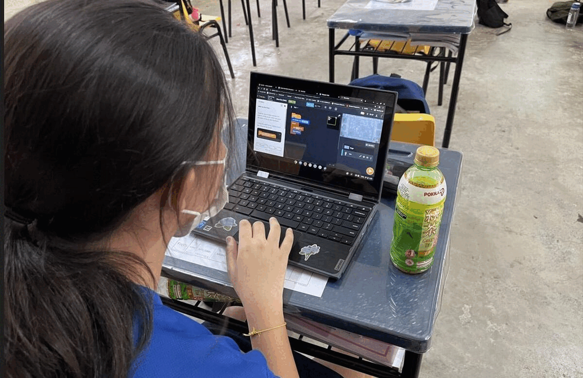A masked girl in a blue shirt uses a laptop showing a coding program at a school desk with a Pokka green tea bottle.